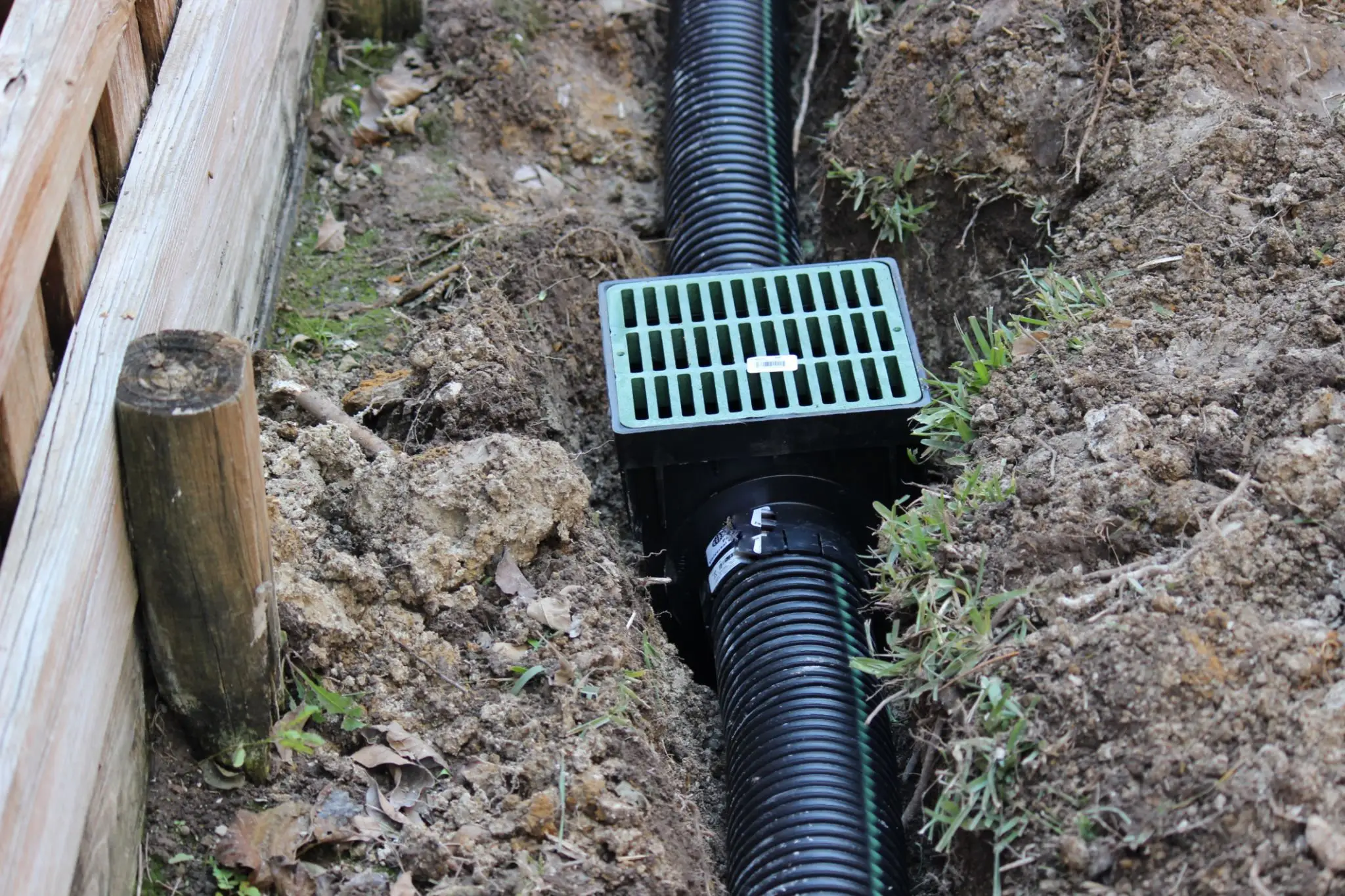 Black drainage pipe with green grate in a muddy trench.